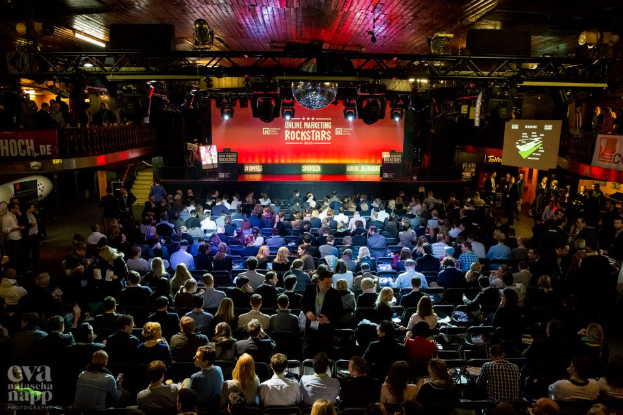 Eine große Menschenmenge sitzt auf Stühlen vor einer Bühne, die von Scheinwerfern beleuchtet und von Geländern umgeben ist. Auf der Bühne befinden sich Bildschirme und Banner mit Text, und in der linken unteren Ecke befindet sich ein Wasserzeichen, was eine Atmosphäre der Aufregung und Vorfreude für ein Online-Marketing-Rockstars-Event schafft.