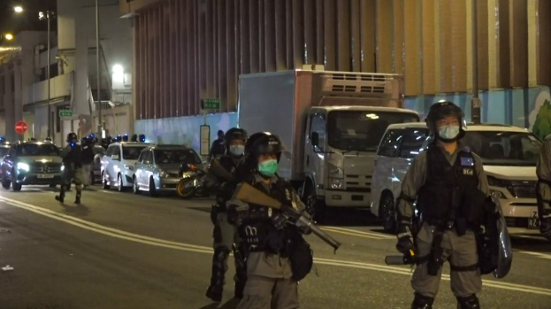 Eine Gruppe von Polizisten in Helmen, Handschuhen und Gesichtsmasken steht auf einer Straße in der Nacht, einige halten Waffen, mit Fahrzeugen, Laternen, Schildern und Gebäuden im Hintergrund.