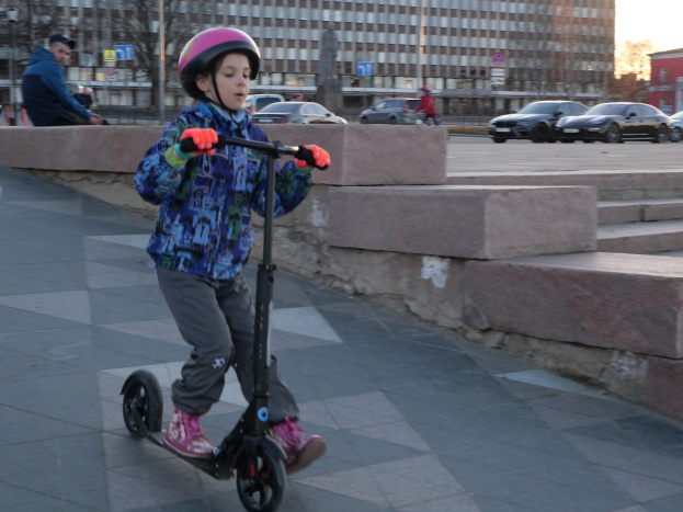 Ein junger Junge in Helm und Handschuhen fährt mit einem Roller eine Treppe hinunter, mit Autos, Menschen, Bäumen, Pfählen, Brettern, Gebäuden und einem klaren blauen Himmel im Hintergrund.