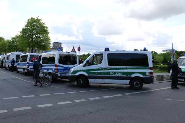 Eine Gruppe von Polizeiwagen steht entlang einer Straße mit einem Radfahrer im Vordergrund, Bäume und Gebäude im Hintergrund unter einem bewölkten Himmel.