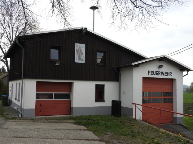 Rotes und weißes Feuerwachengebäude mit einer Straße, Gras, gelben Blumen und Bäumen im Vordergrund und dem Himmel im Hintergrund.