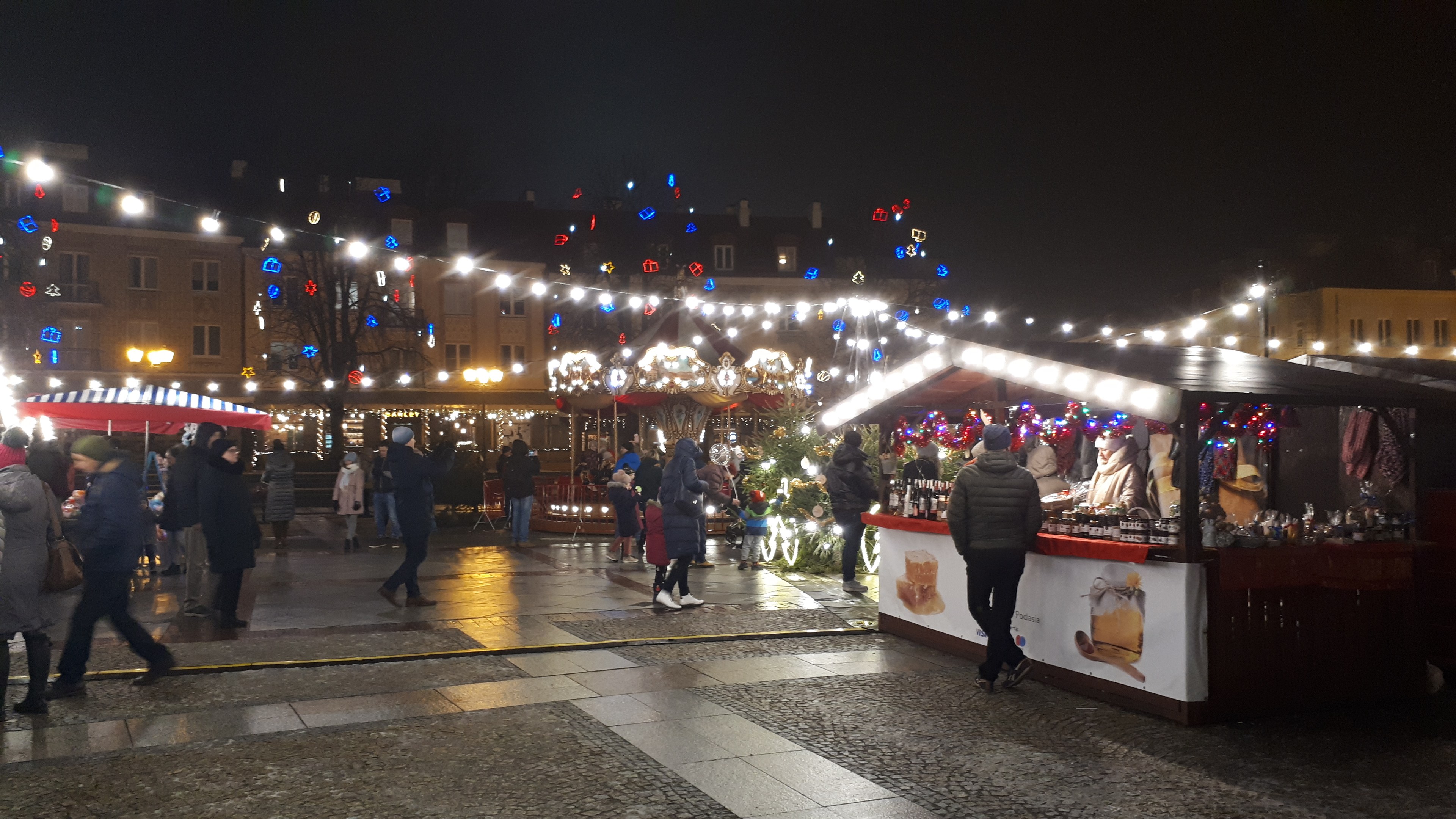 Ein belebter Weihnachtsmarkt bei Nacht in einer Stadt mit Menschen, geschmückten Ständen, beleuchteten Gebäuden, Bäumen und festlicher Beleuchtung am Himmel.
