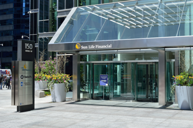 Moderner Glasbau der Sun Life Financial in Toronto mit einer Hinweistafel, Topfpflanzen mit Blüten, Passanten, einer Straßenlaterne, einer Hinweistafel, Fahrzeugen auf der Straße und einem bewölkten Himmel.