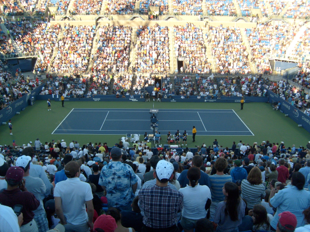 Eine große Menge schaut einem Tennismatch in einem vollen Stadion zu, mit Spielern auf dem Platz und Zuschauern in den Rängen.