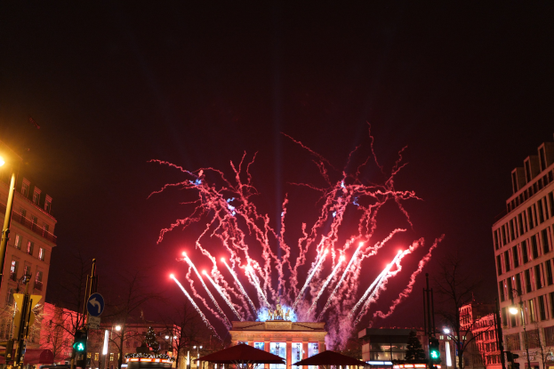 Eine belebte Stadtstraße am Neujahrstag in Berlin, voller Menschen, Fahrzeuge und Gebäude, beleuchtet von Lichtern und Feuerwerk am Himmel.