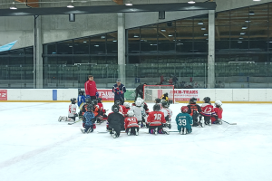 Eine Gruppe von Kindern in Helmen und mit Hockey-Schläger sitzt auf einer Eisbahn, mit einer Wand aus Glas und Säulen, Deckenleuchten und Texttafeln im Hintergrund.