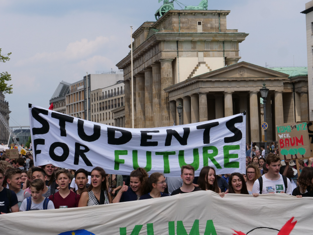 Gruppe von Studenten marschiert in Berlin mit einem bunt bemalten "Students for Future"-Schild vor Gebäuden, Bäumen und Himmel.