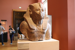 Ein Büste eines ägyptischen Pharaos wird in einem Museum mit einer Gruppe von Menschen, einer Texttafel, einer Frau mit einer Tasche, einer Tür, einer Wand und einer Decke mit Lampen gezeigt.