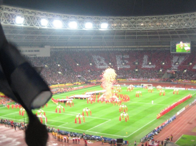 Ein Stadion mit Flutlicht, Eisengittern, Zuschauern, stehenden Personen, Werbetafeln, Bildschirmen, einer Sicherheitskamera, Fahnen und Dekoelementen.