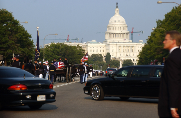 Trauerzug mit Menschen in Mützen und Fahnen vor dem Kapitolsgebäude, begleitet von Fahrzeugen, Laternenmasten, Bäumen und Gebäuden unter einem klaren Himmel.