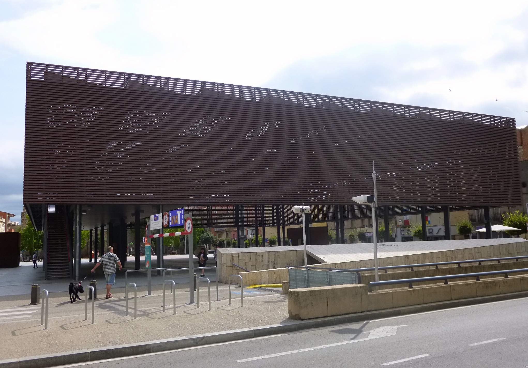Großes Gebäude mit zahlreichen Fenstern, das als neuer Eingang zur Stadtmitte dient, umgeben von Straßeninfrastruktur, Fahrzeugen, Fußgängern und Bäumen unter einem bewölkten Himmel.