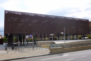 Großes Gebäude mit zahlreichen Fenstern, das als neuer Eingang zur Stadtmitte dient, umgeben von Straßeninfrastruktur, Fahrzeugen, Fußgängern und Bäumen unter einem bewölkten Himmel.