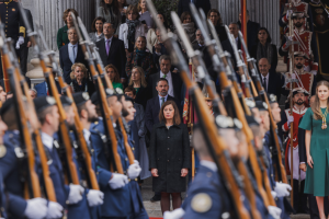 Eine Gruppe von Menschen in militärischer Kleidung steht vor einer Menge, einige halten Gewehre, während sie an Säulen und Grün vorbeimarschieren.