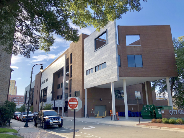 Großes mehrstöckiges Stadt Hall Apartments-Gebäude mit zahlreichen Fenstern, Straßeninfrastruktur einschließlich Pfosten, Lampen und Verkehrszeichen, umgeben von Fahrzeugen, Grünflächen und einem bewölkten Himmel.