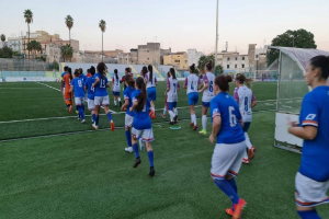 Eine Gruppe von Frauen, die Fußball auf einem Feld spielen, das von Gebäuden, Bäumen und Pfählen umgeben ist, unter einem klaren blauen Himmel, mit einem Torpfosten und Netz auf der rechten Seite.
