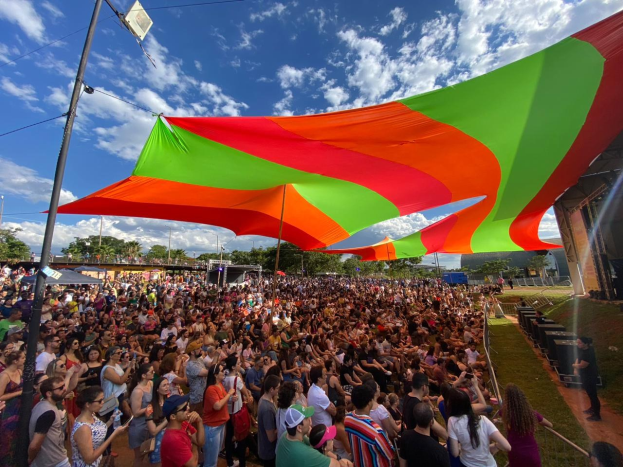 Eine große Menschenmenge steht vor einer Regenbogenflagge, mit einem Gebäude, einer Bühne, Bäumen und Wolken im Hintergrund, die sich zu einem Festivaljubiläum versammelt hat.