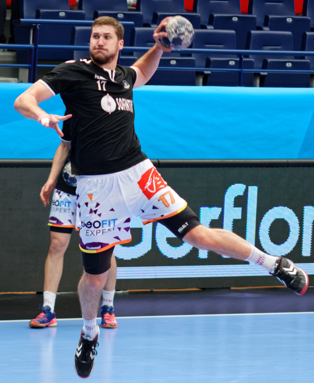 Ein Mann in schwarzem T-Shirt und weißen Shorts hält einen Handball in einem Stadion mit Zuschauern im Hintergrund.