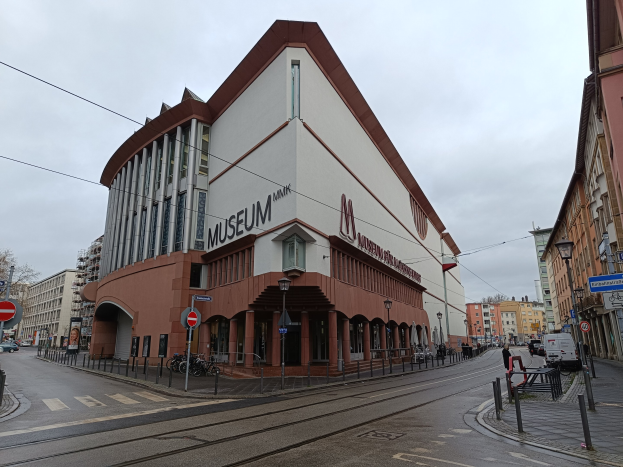 Großes Gebäude mit vielen Fenstern, identifiziert als Museum of Modern Art in München, Deutschland, umgeben von Straßeninfrastruktur, Fahrzeugen, Fußgängern, Bäumen und einem bewölkten Himmel.