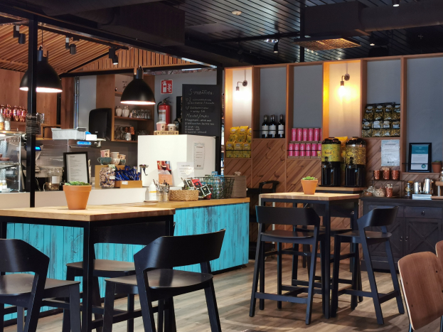 Innenraum eines Restaurants mit Holztischen und -stühlen, einem Blumentopf auf einem Tisch, einer Schiefertafel, einem Kühlschrank, einem Feuerlöscher, Regalen mit Flaschen, Schränken mit Gegenständen, einem gerahmten Foto an der Wand und Deckenleuchten.