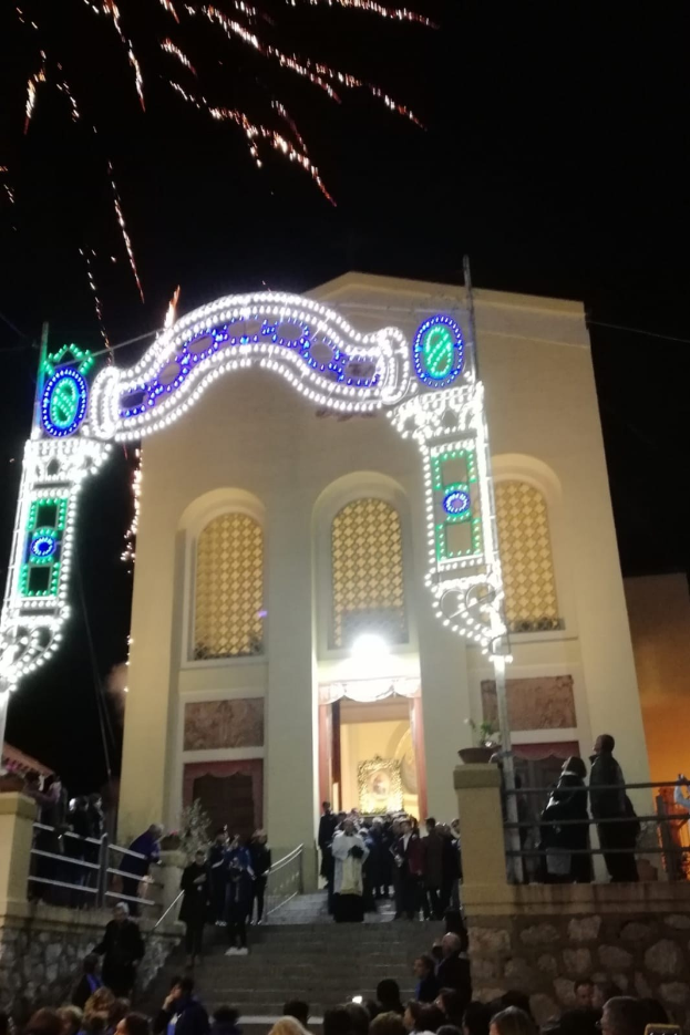 Eine Gruppe von Menschen steht vor einem Gebäude mit einem beleuchteten Bogen, Treppen mit Geländern und Topfpflanzen, während farbenfrohe Raketen den Nachthimmel erhellen.