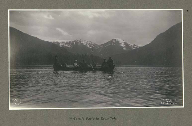 Schwarzes und weißes Foto einer Familie in einem Kanu auf einem See mit Bergen und Bäumen im Hintergrund, Himmel sichtbar und Text unten.