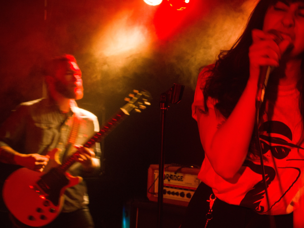 Eine Frau singt in ein Mikrofon auf der Bühne, während ein Mann Gitarre spielt und Deckenlampen das lebhafte Setting beleuchten.