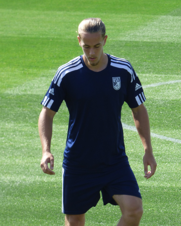 Ein professioneller Fußballspieler in einem blauen T-Shirt und Shorts geht selbstbewusst über ein saftiges grünes Feld.