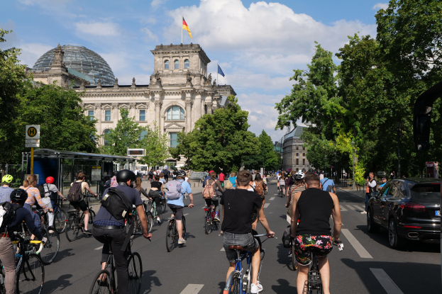 Eine Gruppe von Menschen, die auf Fahrrädern eine von Bäumen gesäumte Straße in Berlin, Deutschland, entlangfahren, mit Gebäuden auf der linken Seite und einer Bushaltestelle auf der rechten Seite.
