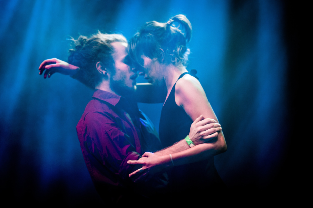 Zwei Menschen, die sich auf einer Bühne umarmen und küssen, vor dunklem Hintergrund.
