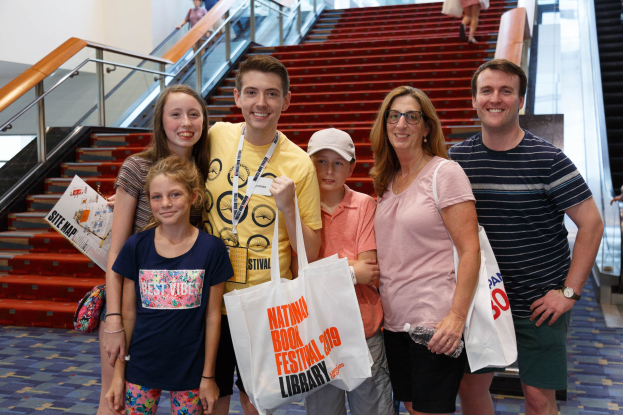 Gruppe von Menschen, die vor einer Rolltreppe im Nationalen Buchfestival 2019 lächeln und Taschen halten, mit Treppen und Geländern im Hintergrund.
