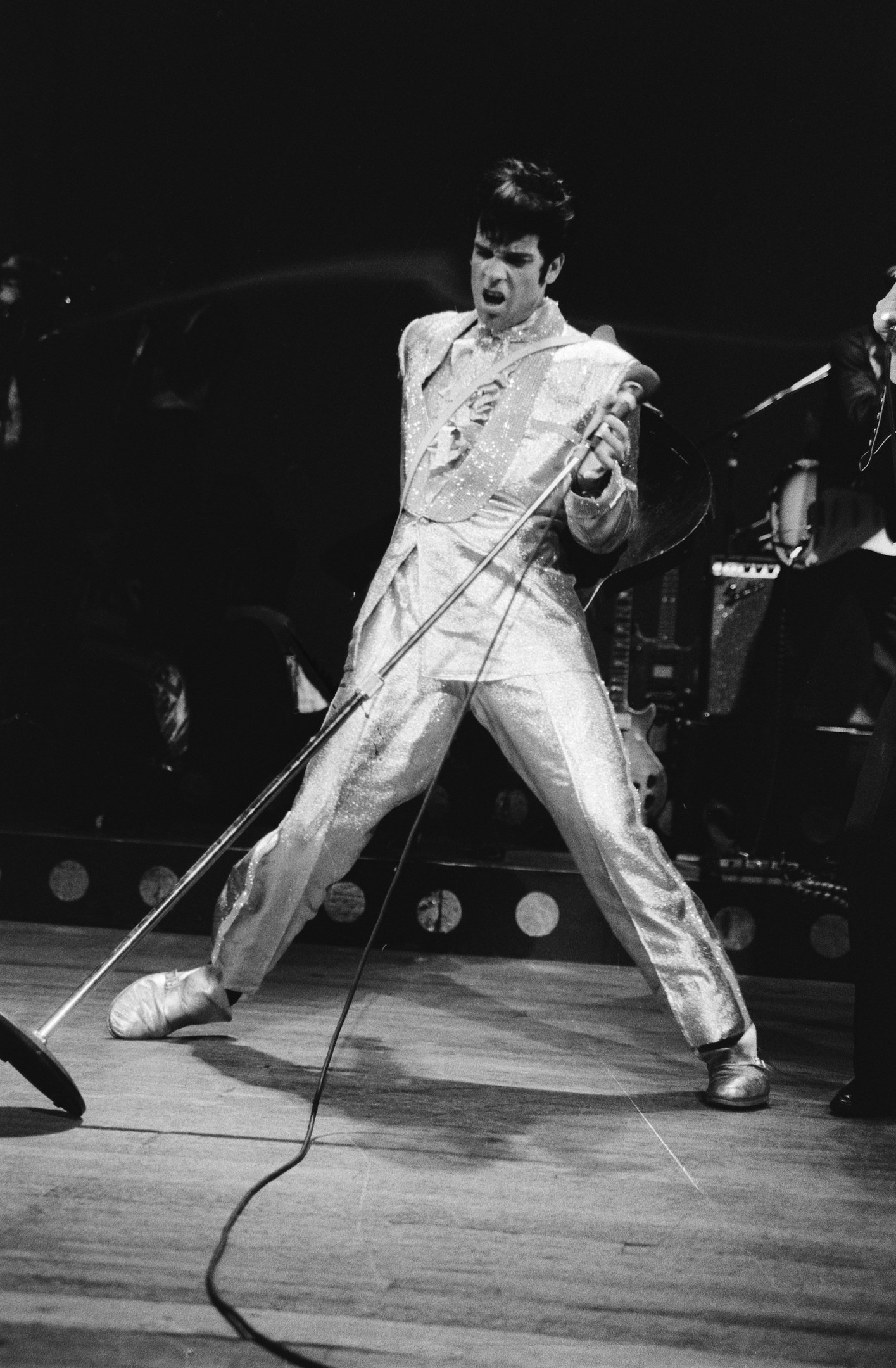 Elvis Presley in weißer Hose performt auf der Bühne im Apollo Theater in Las Vegas, hält ein Mikrofon in der Hand, während ein anderer Musiker rechts von ihm mit einem Instrument steht; Bühnenlichter und Musikinstrumente sind im Hintergrund zu sehen.