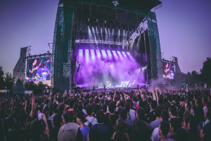 Eine große Menschenmenge auf einem Musikfestival, mit Performern auf einer zentralen Bühne, die von Scheinwerfern beleuchtet und von Bildschirmen umgeben ist, unter einem klaren blauen Himmel mit einem Gebäude und Bäumen im Hintergrund.