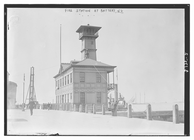 Schwarzes und weißes Foto einer Feuerwache an der Batterie in New York mit einem Gebäude, Menschen, Pfählen, einem Zaun, Booten auf dem Wasser und einem bewölkten Himmel.