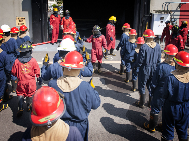 Feuerwehrleute in roter Uniform und Helmen gehen eine Straße entlang an einer Wand mit Schrift vorbei, mit Bäumen und einem klaren blauen Himmel im Hintergrund.