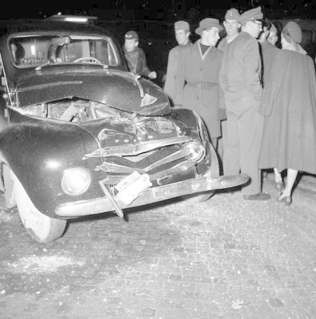 Schwarz-weißes Bild eines schwer beschädigten Autos am Straßenrand mit eingedellter Motorhaube und zersplitterten Fenstern, umgeben von einer Gruppe von Menschen, die die Szene begutachten.