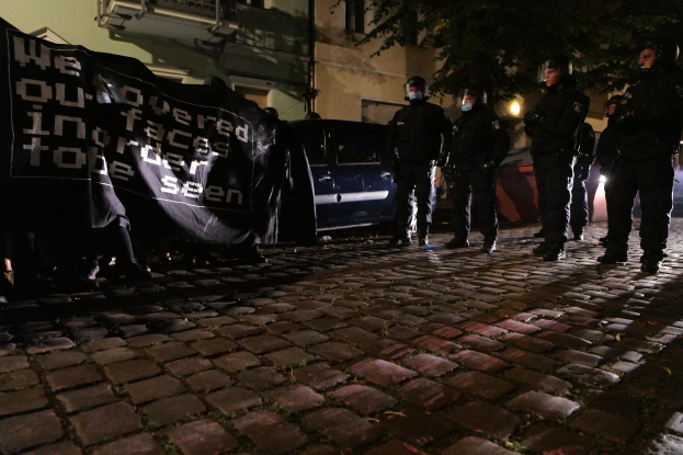 Gruppe von Polizisten mit Helmen, die ein Banner auf einer Kopfsteinpflasterstraße bei Nacht halten, mit Fahrzeugen, Gebäuden, Bäumen und Straßenbeleuchtung im Hintergrund.