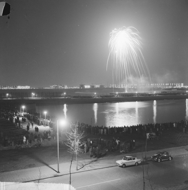 Ein Schwarz-Weiß-Foto von einer Menschenmenge, die Feuerwerke über einem Gewässer beobachtet, mit Fahrzeugen auf der Straße, Laternenpfählen, Bäumen und Gebäuden im Hintergrund.